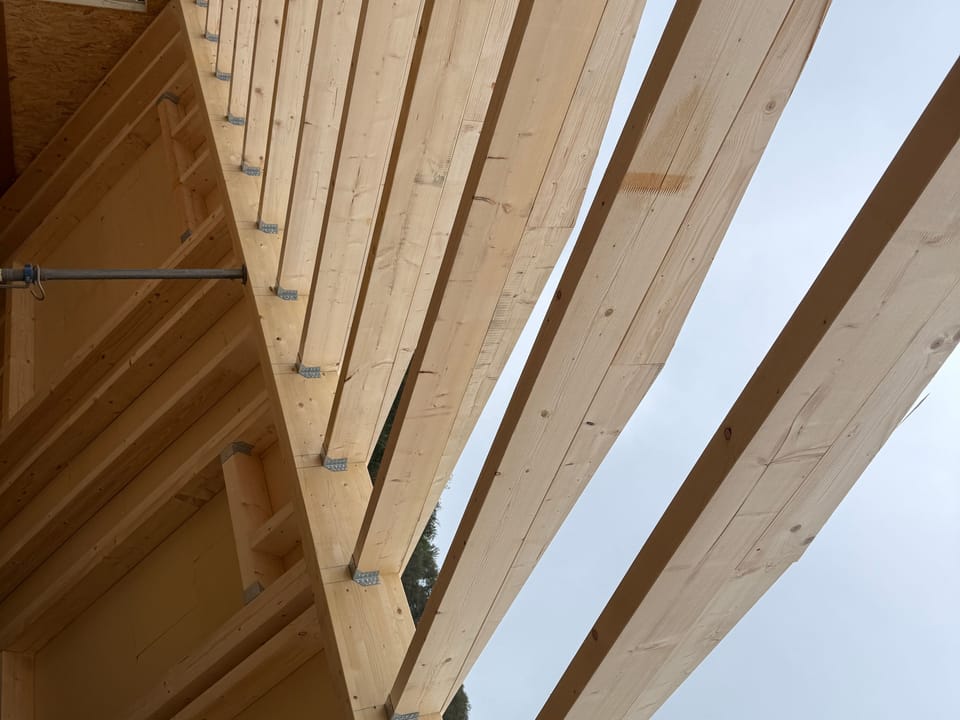 Blick von unten auf neue Holzsparren, die mit Stahlwinkeln auf einer Holzrahmenwand befestigt sind, dahinter grauer Himmel