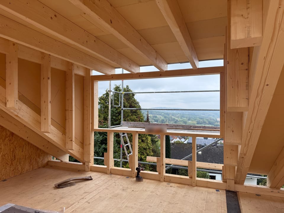 Blick aus dem neuen Geschoss in Holzbauweise: Holzbalkendecke, Holzrahmenwände und eine große Fensteröffnung mit Blick über Dächer und Gerüst ins Tal