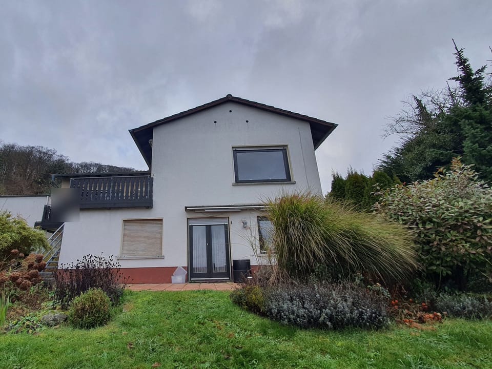 Weiß verputztes Einfamilienhaus mit Satteldach von der Gartenseite, Balkon mit dunklem Holzgeländer, davor Rasen und Sträucher